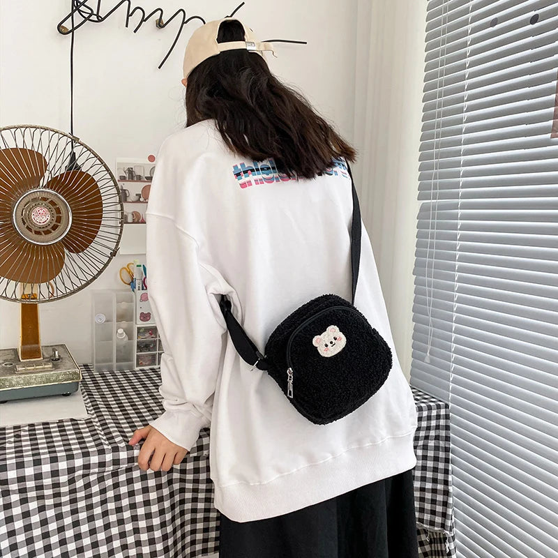 Person wearing a white sweatshirt with text and a black Kawaii bear-themed bag, standing in a room with a fan and window blinds.