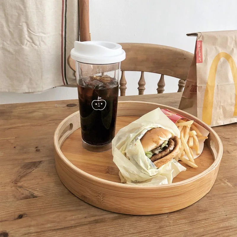 Burger, fries, and a Kawaii-styled coffee cup on a wooden tray with a chair and bag in the background.
