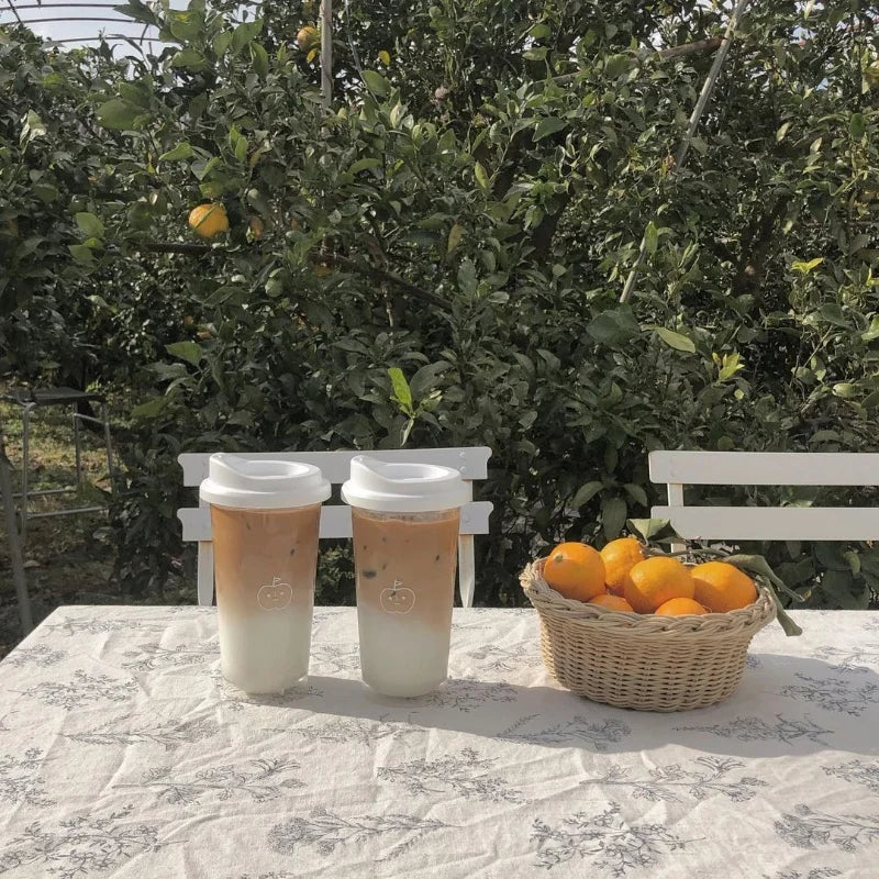 Two Kawaii coffee cups with a white lid on a table outdoors, with a basket of oranges and greenery in the background.