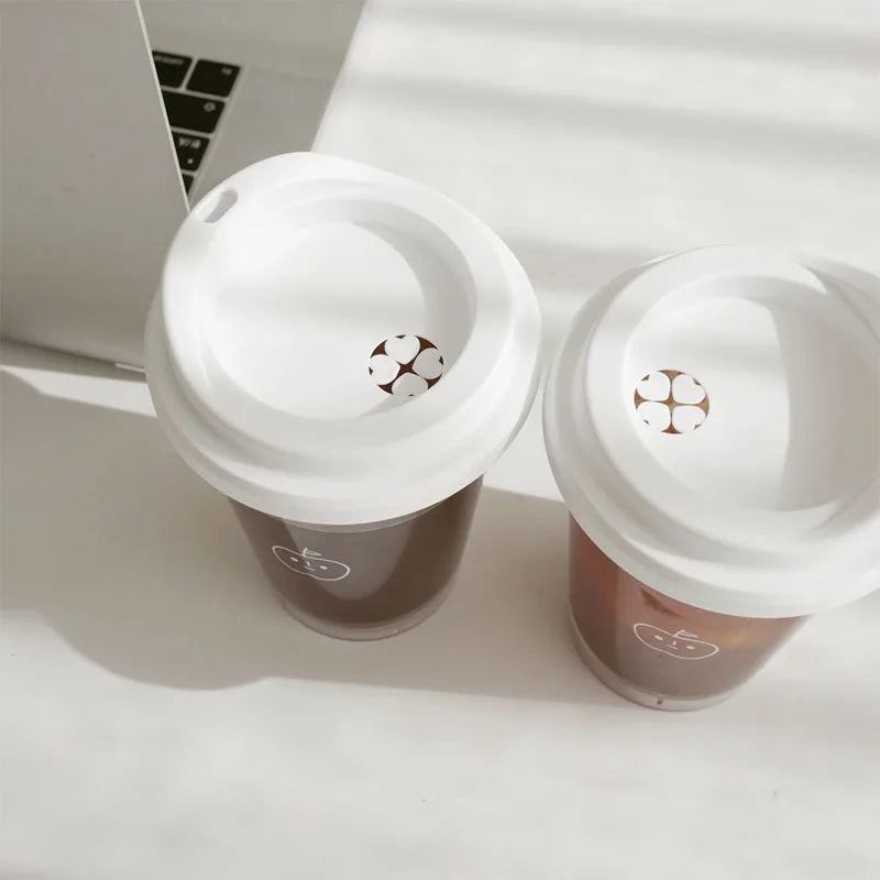 Two Kawaii coffee cups with lids on a white surface next to a laptop.