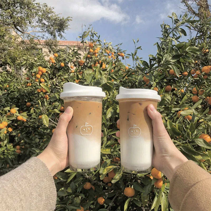 Two hands holding transparent Kawaii-inspired cups with white lids against a backdrop of orange trees.