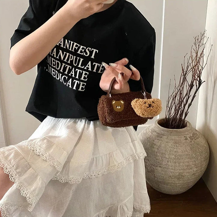 Person holding a small brown handbag with a Kawaii teddy bear design, wearing a black t-shirt with text and a white skirt.