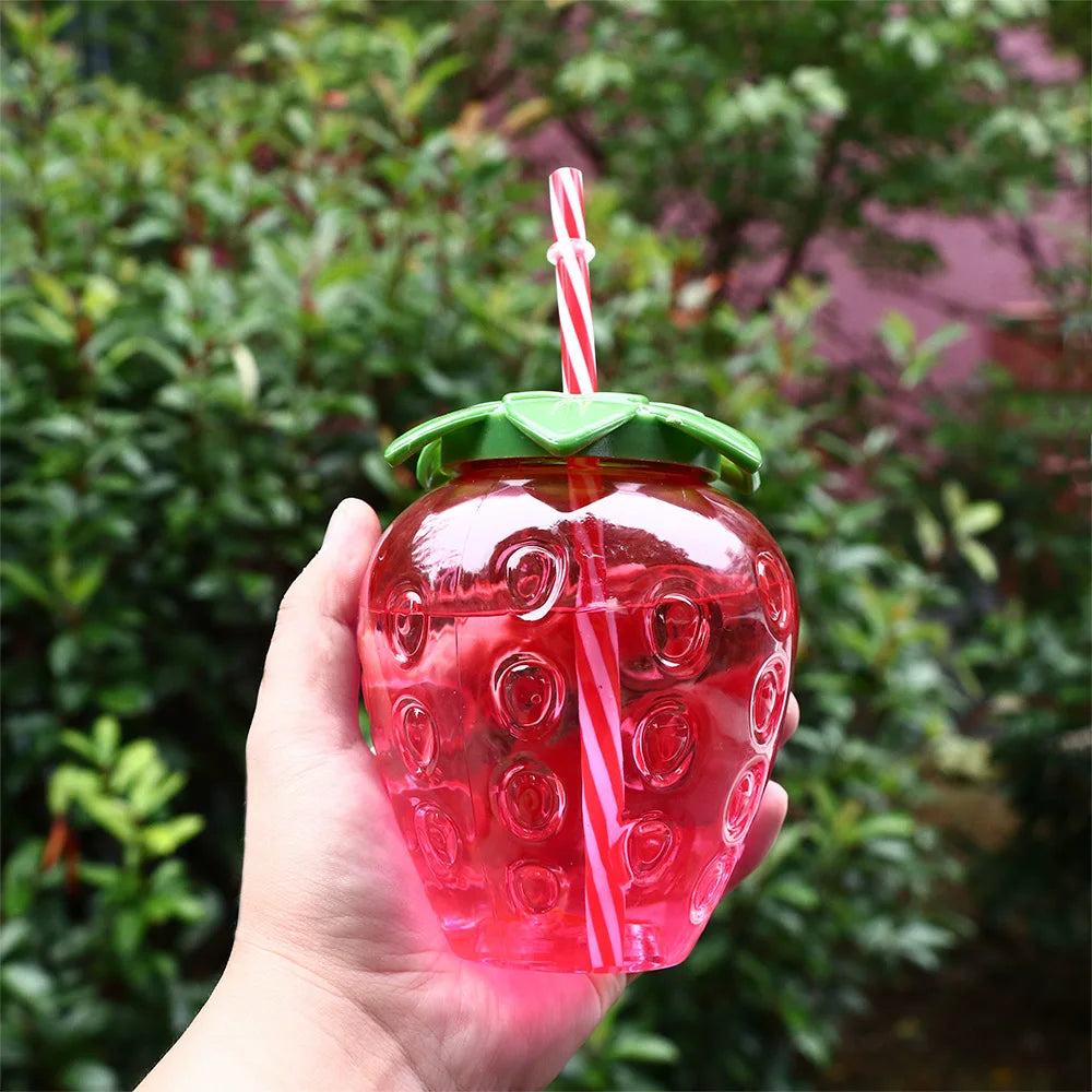 Strawberry-shaped Kawaii tumbler with a green lid and red straw held by a hand against a blurred green background