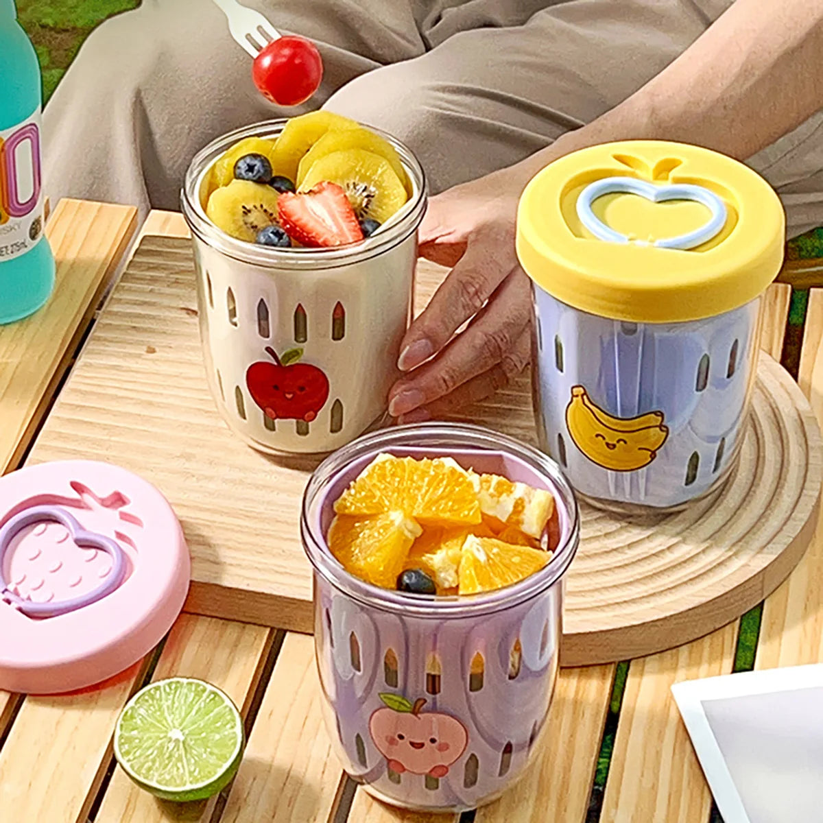 Three transparent Kawaii containers with fruit inside on a wooden table, surrounded by a lime and a bottle.