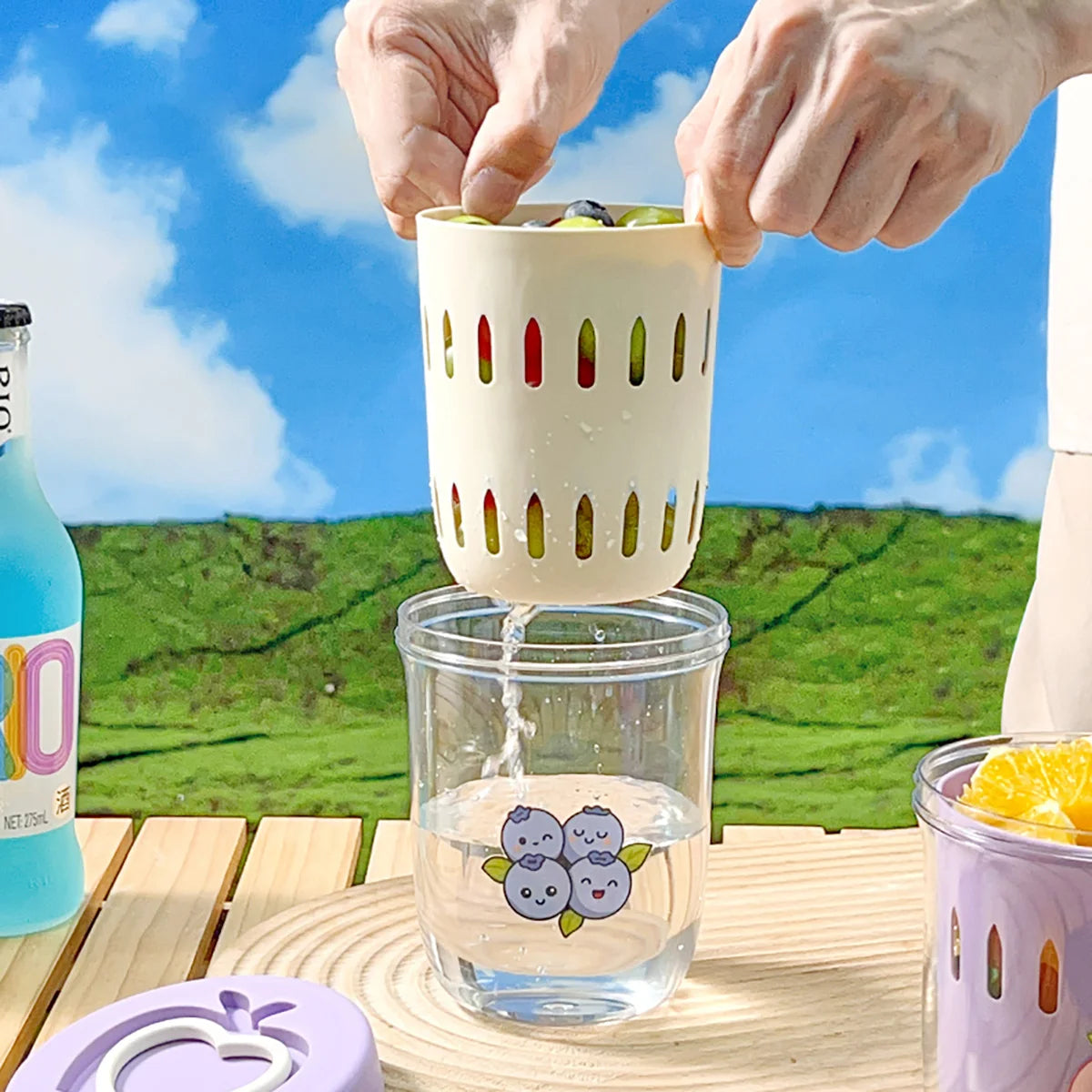 Person using a colorful fruit infuser over a Kawaii-inspired tumbler with a scenic background