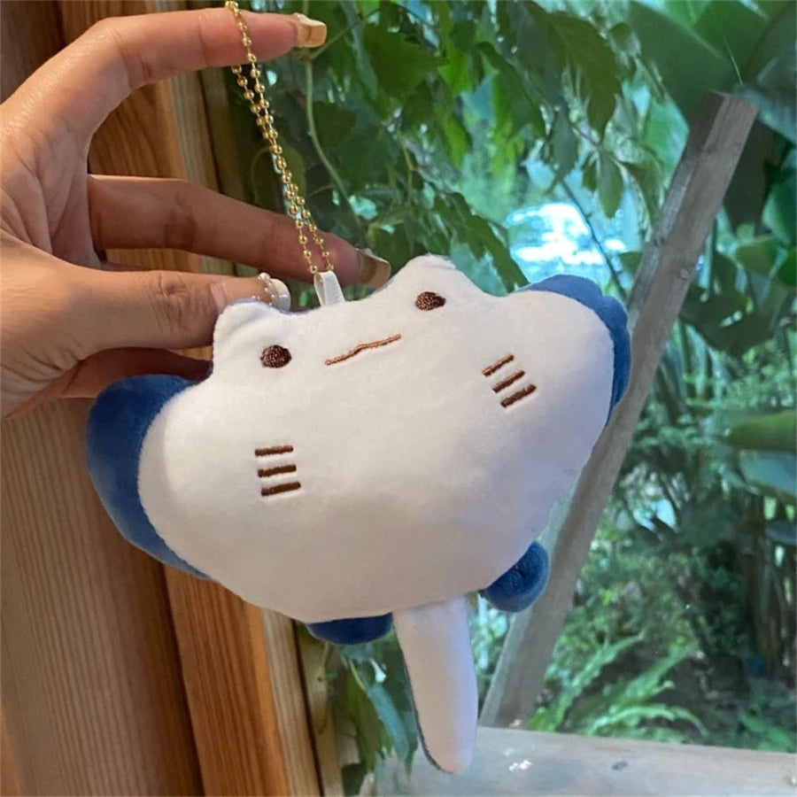 Plush kawaii keychain shaped like a cat with blue ears held by a hand against a blurred green outdoor background.