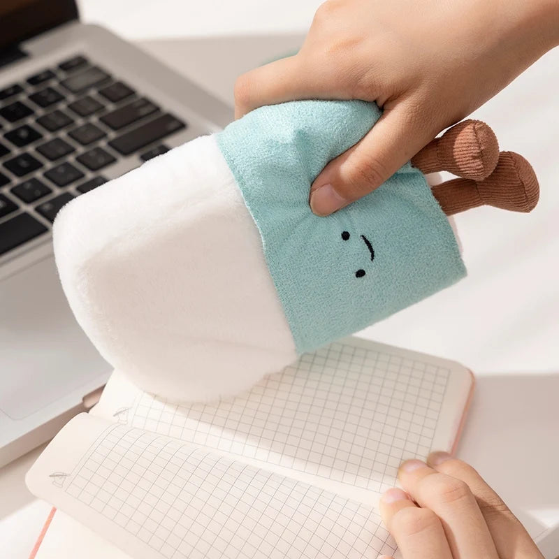 Hand holding a kawaii plush toy resembling a cartoon character next to a laptop and notebook.
