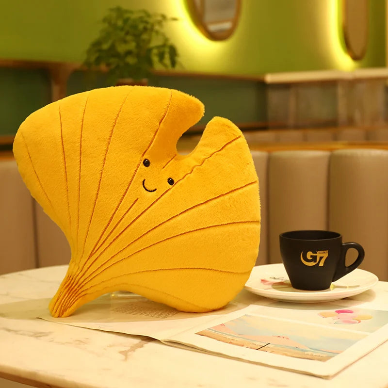 Yellow leaf-shaped kawaii pillow with a smiley face on a table with a coffee cup.