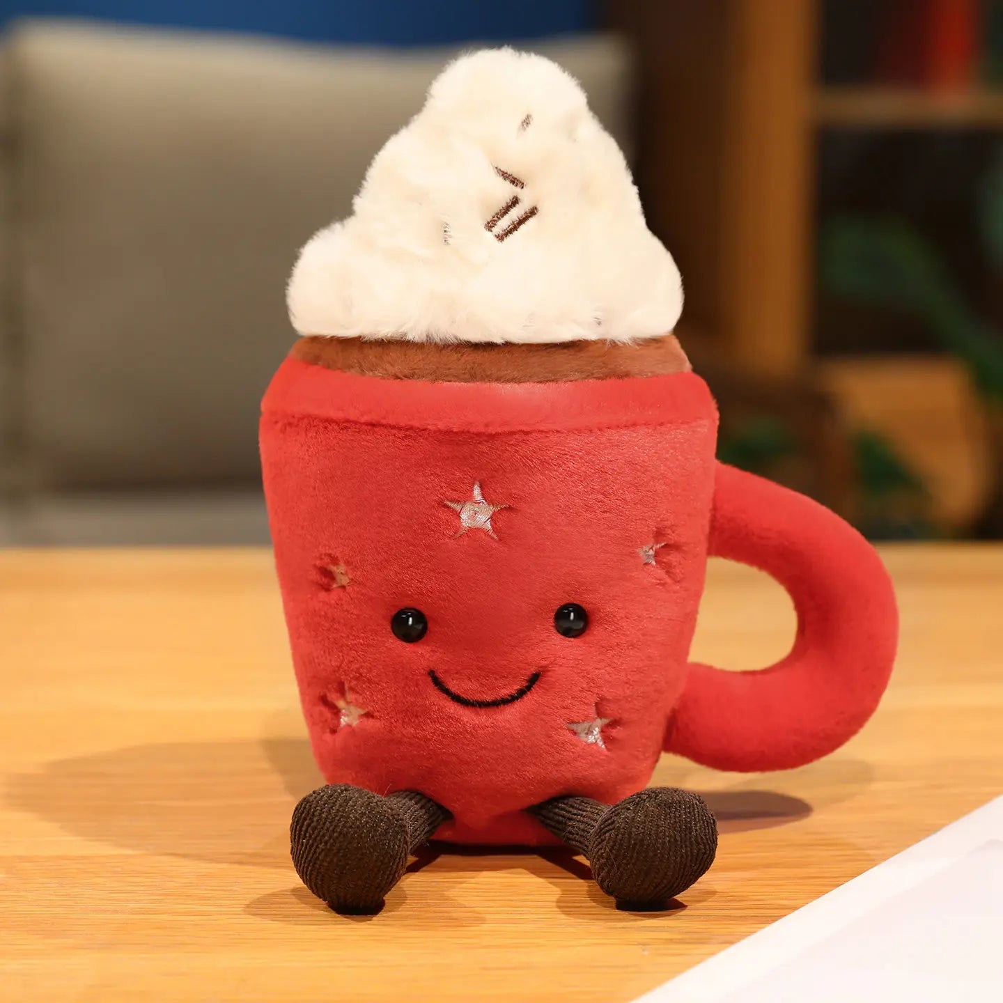 Red kawaii plush mug with a smiling face, whipped cream, and chocolate pieces on a wooden surface.