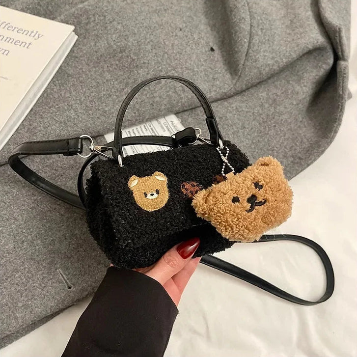 Black handbag with Kawaii teddy bear keychain held by a hand on a gray surface.