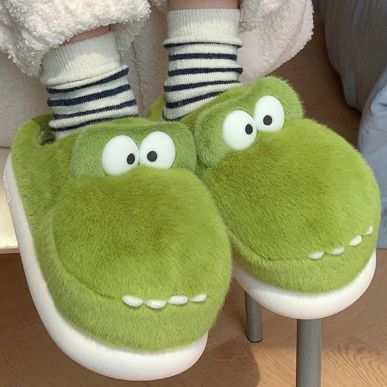 Green kawaii cartoonish slippers with white eyes and a mouth worn by a person.