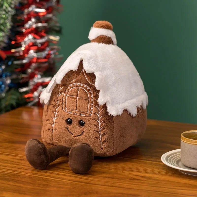 Kawaii plush gingerbread house toy with snow on a wooden table with a Christmas tree in the background.
