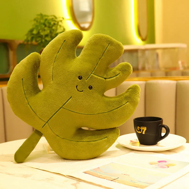 Green plush kawaii toy shaped like a leaf with a face on a table with a cup and saucer.