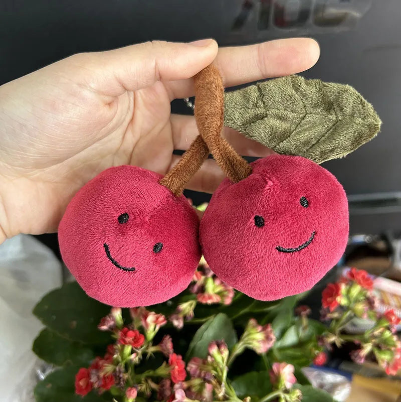 Plush kawaii cherries with smiling faces held by a hand, with a blurred background.