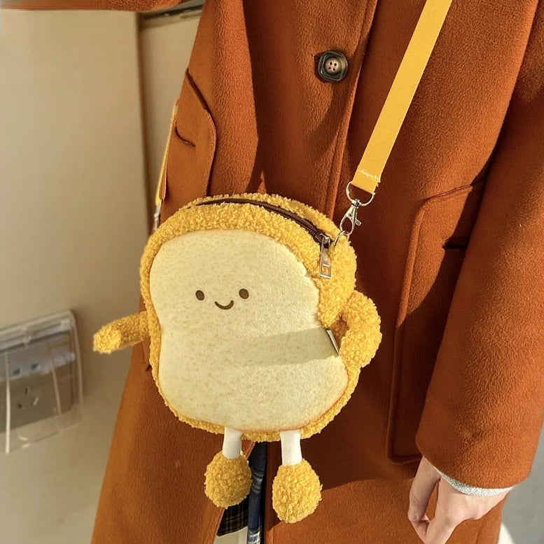 Kawaii plush toy shaped like a slice of bread with arms and legs attached to a yellow strap over a person wearing an orange coat.