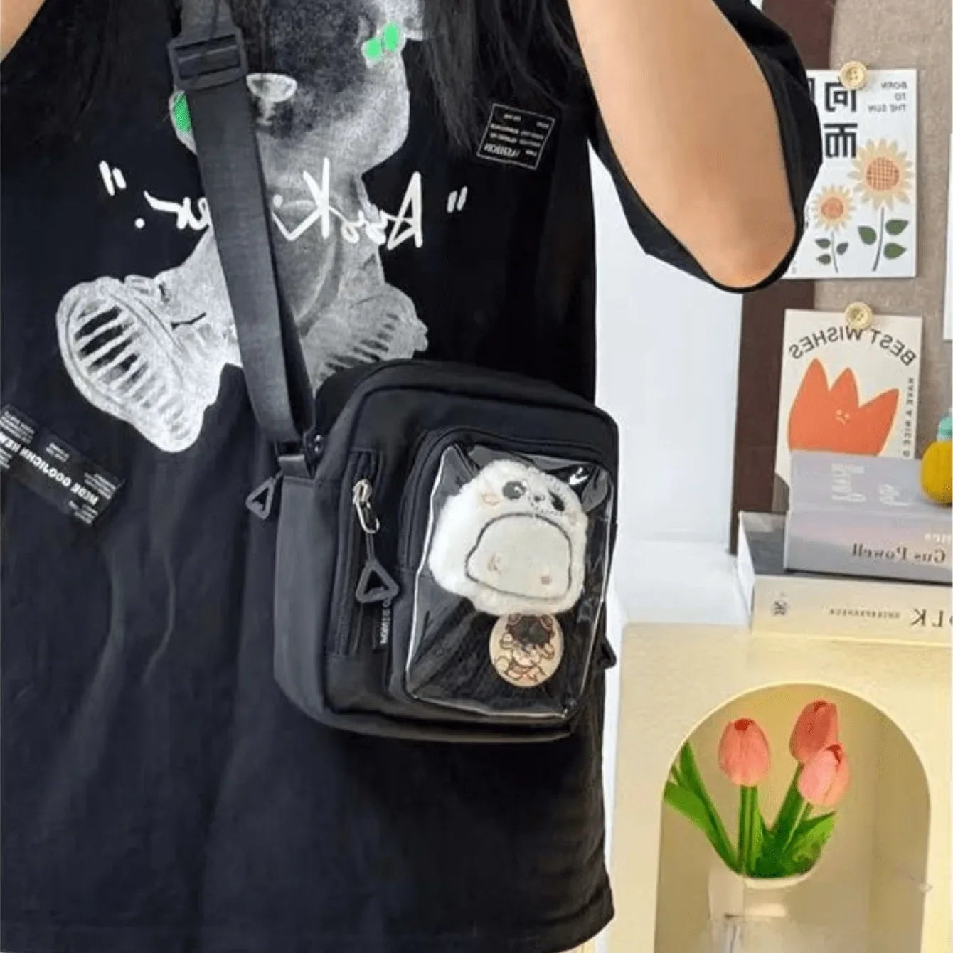Person wearing a black bag with a transparent pocket containing a small Kawaii plush toy, standing next to a display with flowers and cards.