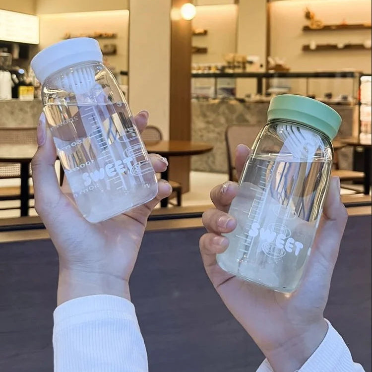 Two hands holding clear Kawaii glass bottles with white labels in a blurred indoor setting.