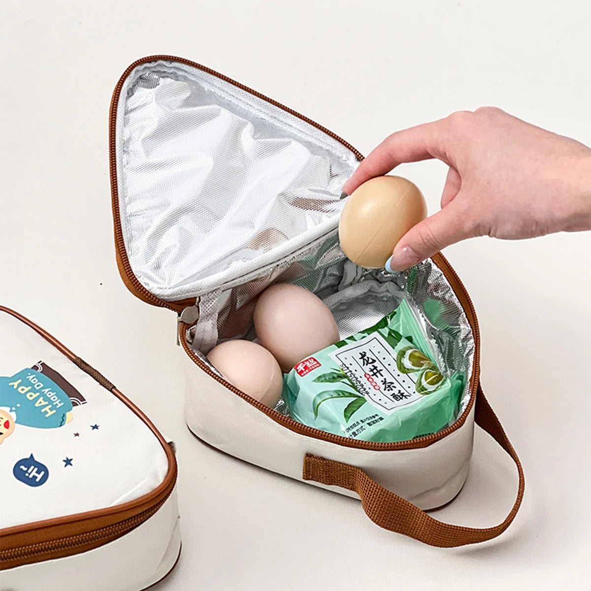 Kawaii-style small insulated food container with eggs and a snack, held by a hand on a light background
