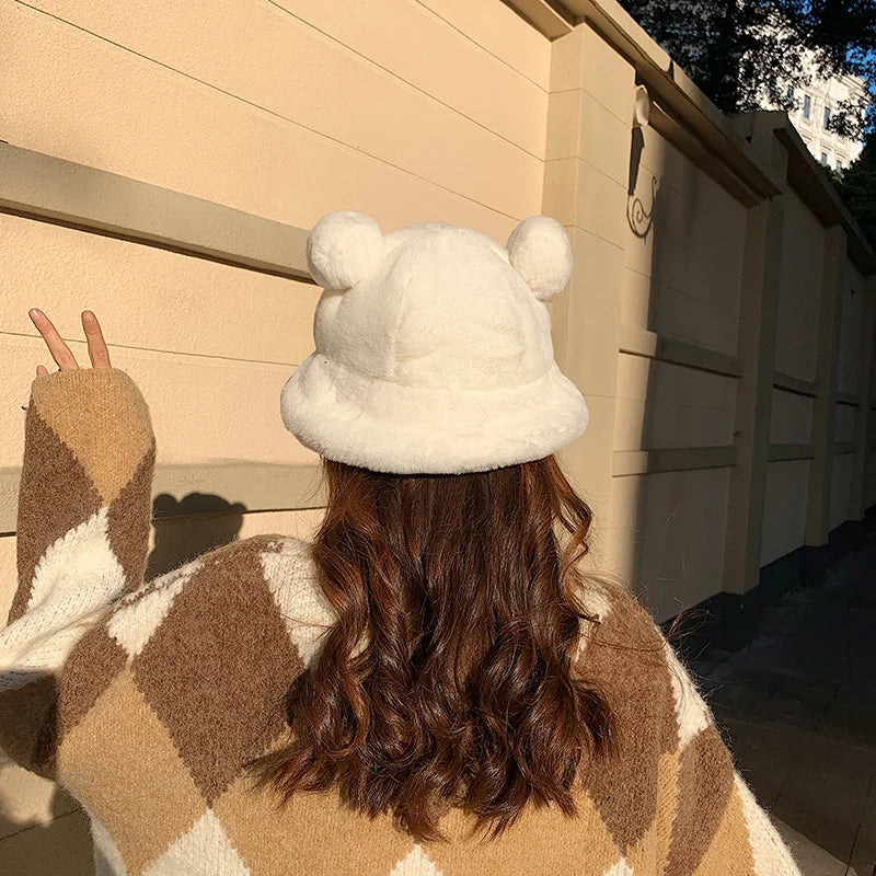 Person wearing a fluffy white Kawaii hat and checkered coat standing in front of a beige wall.
