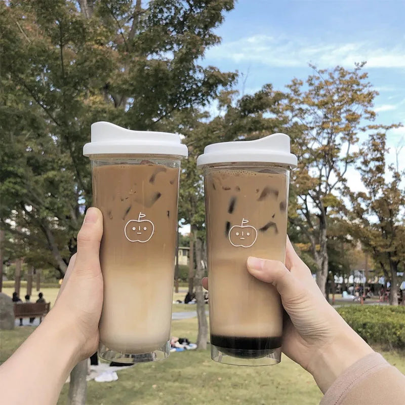 Two hands holding transparent Kawaii cups with white lids, filled with a brown beverage, against a park background.