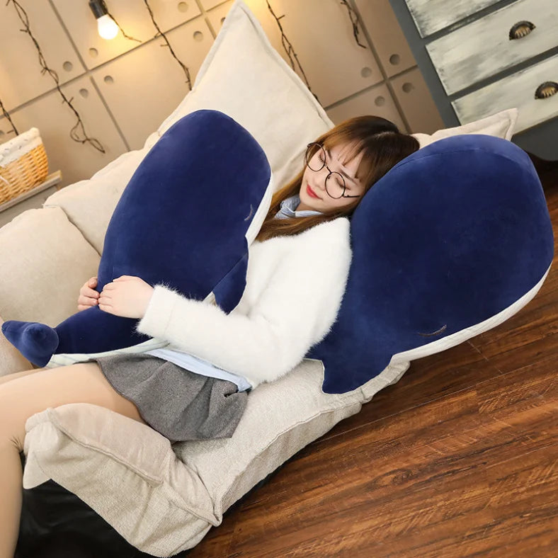 Person lying on a couch with a large blue and white plush kawaii toy shaped like a whale.