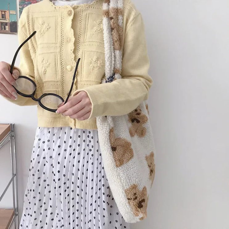 Person wearing a yellow kawaii cardigan holding glasses, with a white wall and wooden shelf in the background