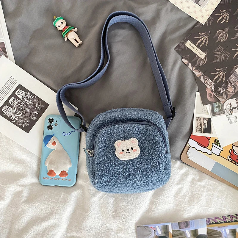 Blue textured crossbody bag with Kawaii bear emblem on a bed with magazines and a phone.