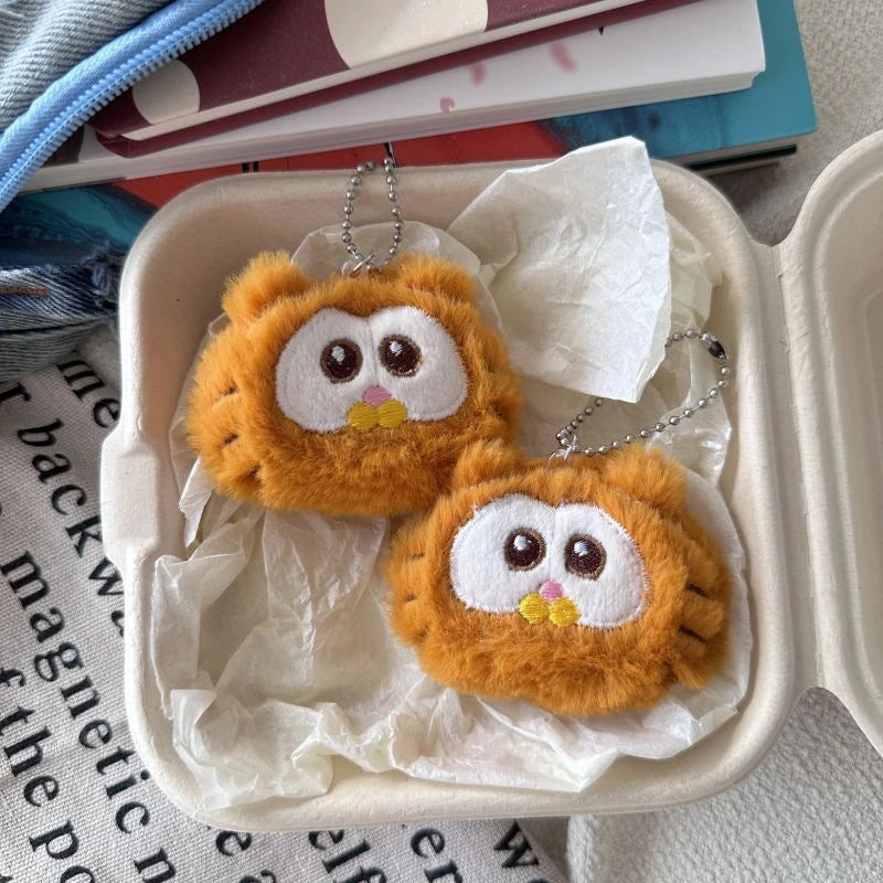 Two kawaii orange plush owl keychains in a takeout container on a newspaper background.