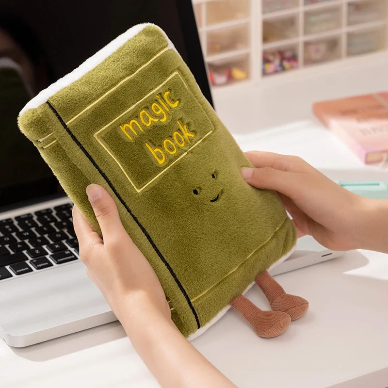 Green kawaii plush toy shaped like a book with 'magic book' text, held by a person in front of a laptop.