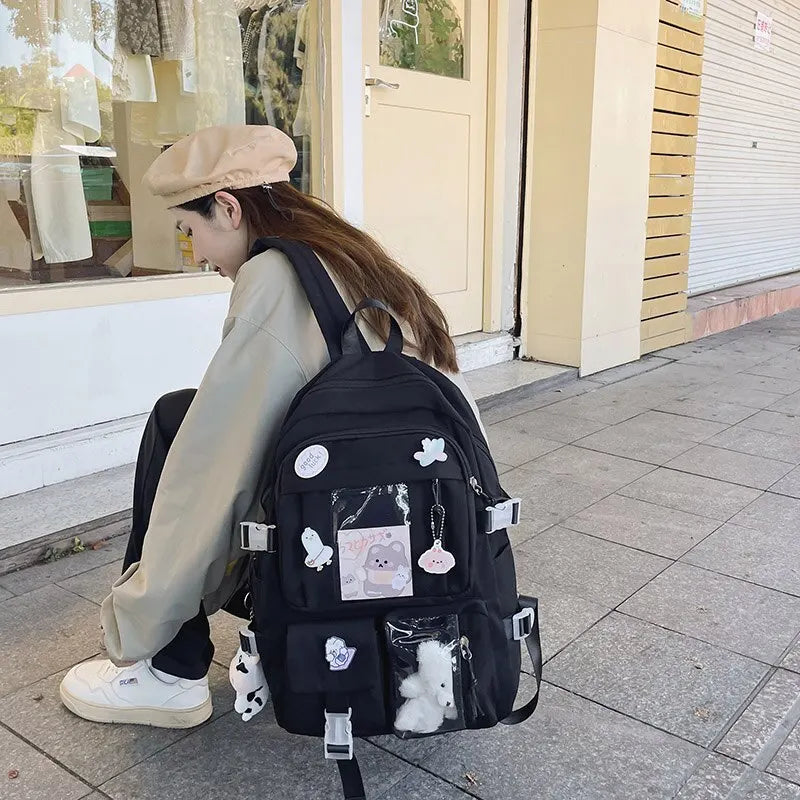 Person sitting on a sidewalk with a black backpack decorated with stickers and a Kawaii small plush toy.