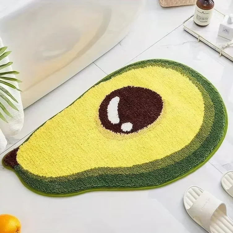 Avocado-shaped Kawaii bath mat on a bathroom floor with a plant and slippers in the background.