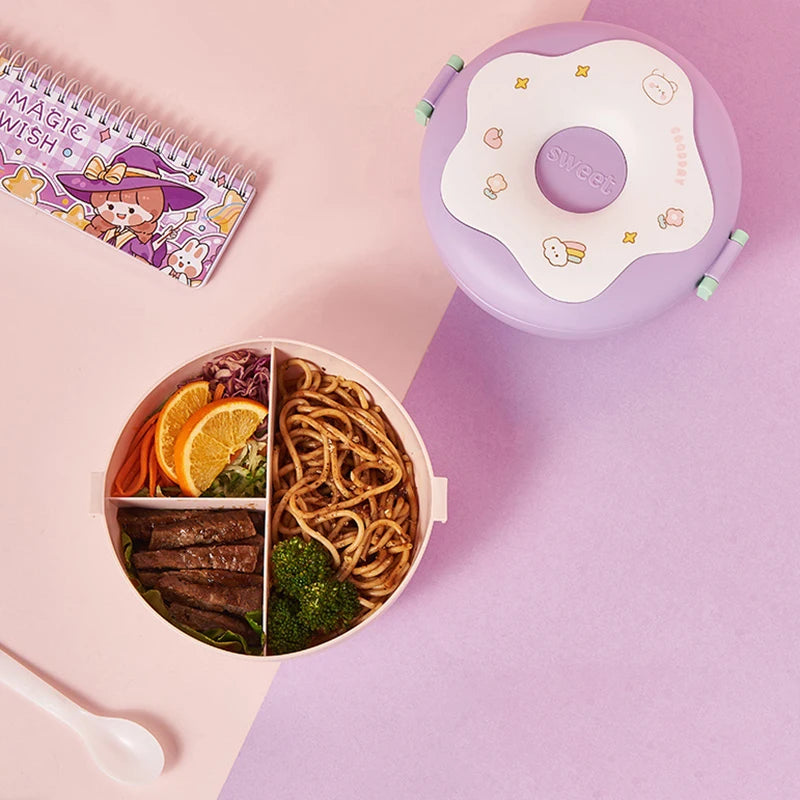 Divided kawaii lunch container with food on a pink surface with a purple lid and 'Magic Wish' notebook.