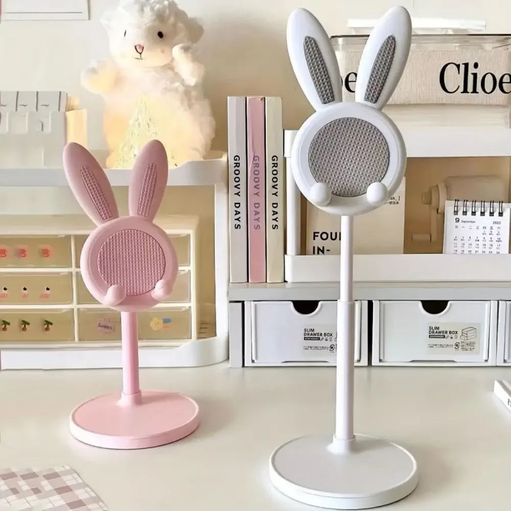 Two kawaii phone stands with bunny ears, one pink and one white, on a surface with books and a teddy bear in the background.