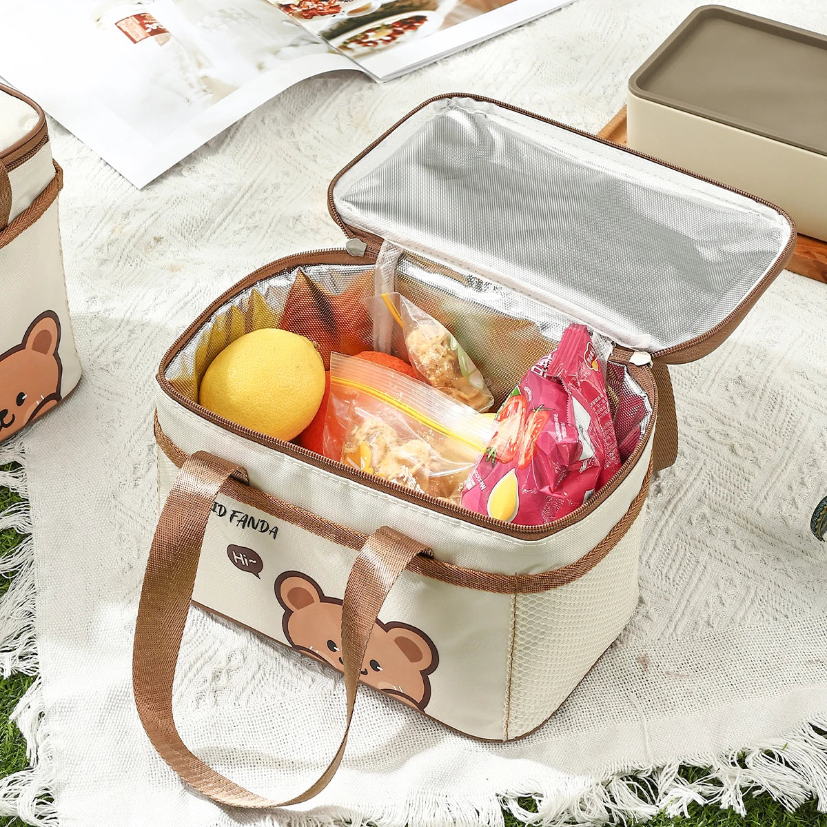 Insulated lunch bag with kawaii cartoon panda design filled with snacks on a picnic blanket.