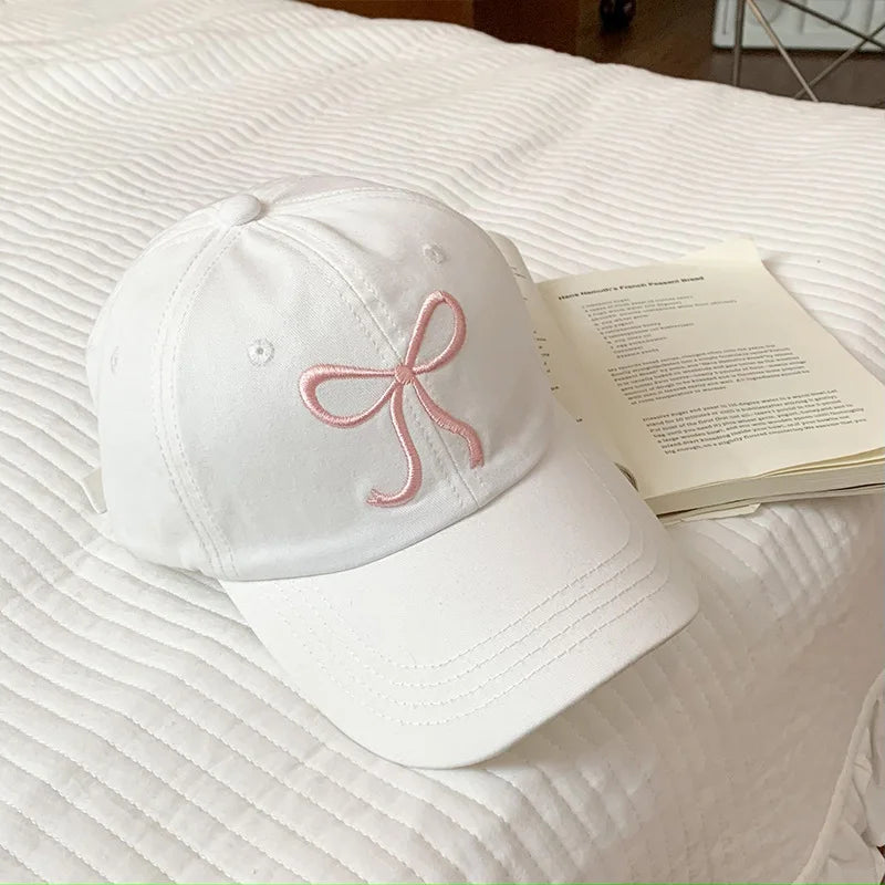 White Kawaii cap with pink bow design on a bed next to an open book