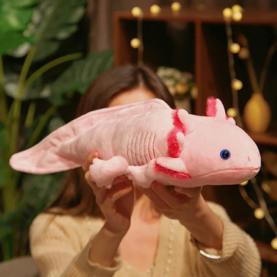 Person holding a pink axolotl kawaii plush toy with red markings in a blurred indoor setting.