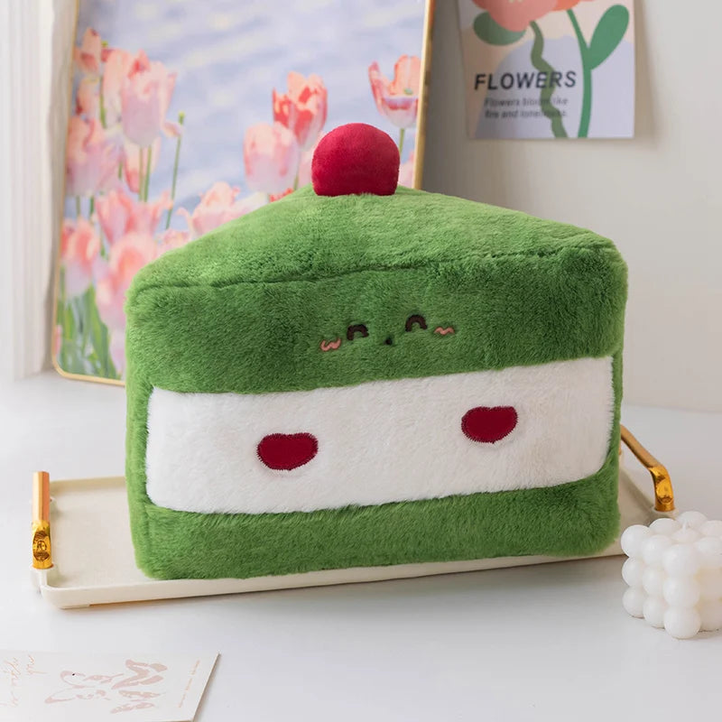 Green and white kawaii plush toy resembling a slice of cake with a red cherry on top, placed on a white surface with a floral painting in the background.