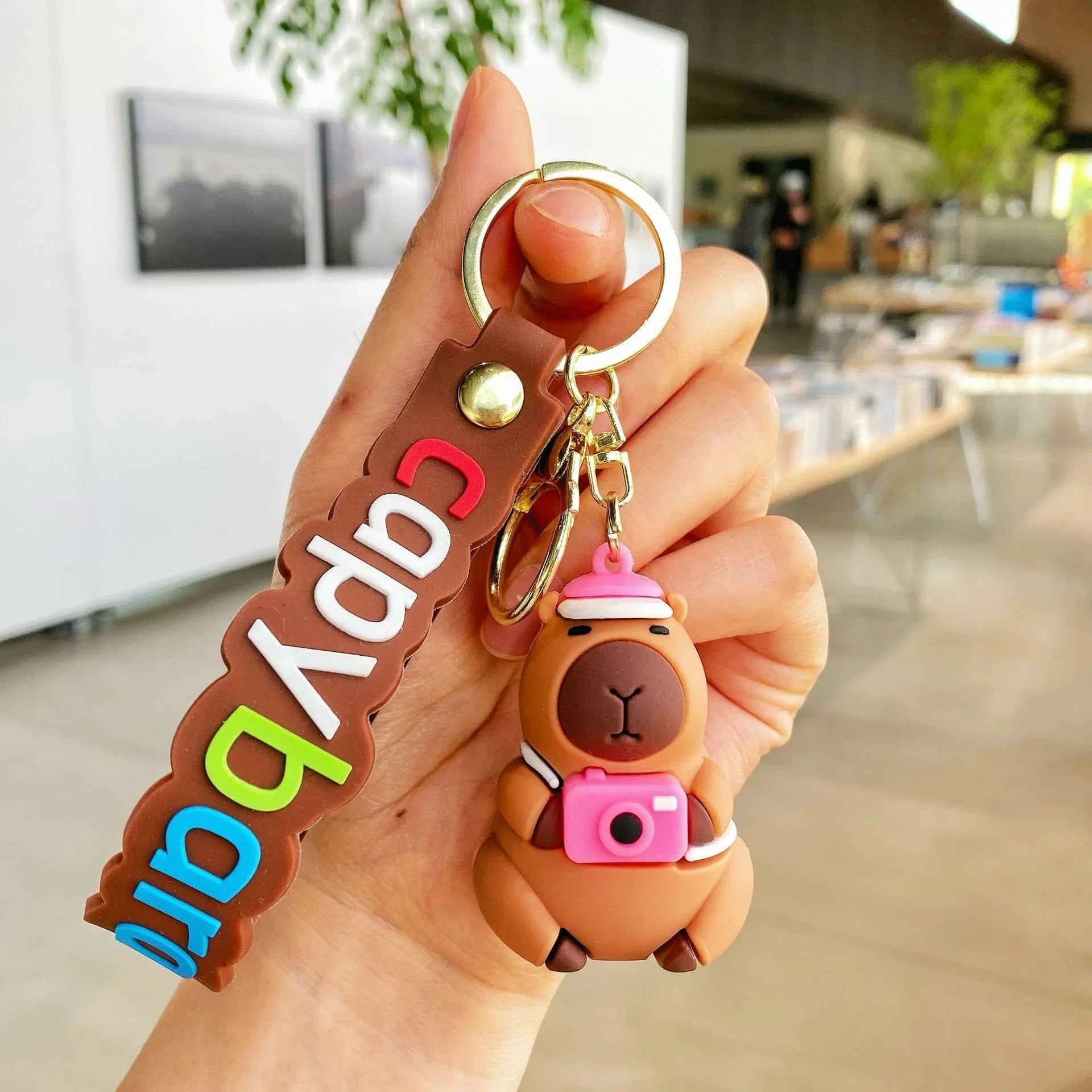 Hand holding a kawaii keychain with a cartoon bear and colorful text on a blurred indoor background