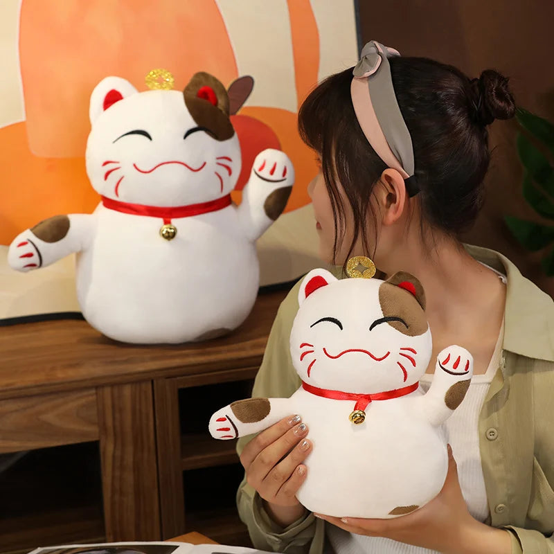 Woman holding a large white cat-shaped kawaii plush toy with a red ribbon and gold bell, sitting next to another similar plush toy on a wooden table.