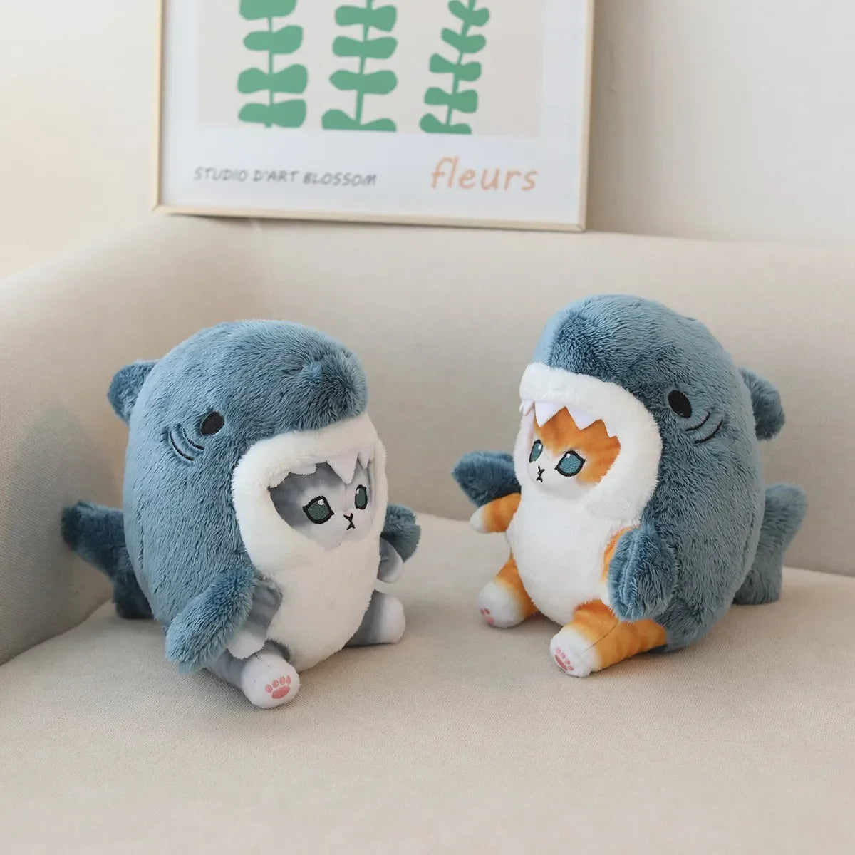 Two kawaii plush toys resembling sharks with cat faces on a beige couch, with a framed picture in the background.