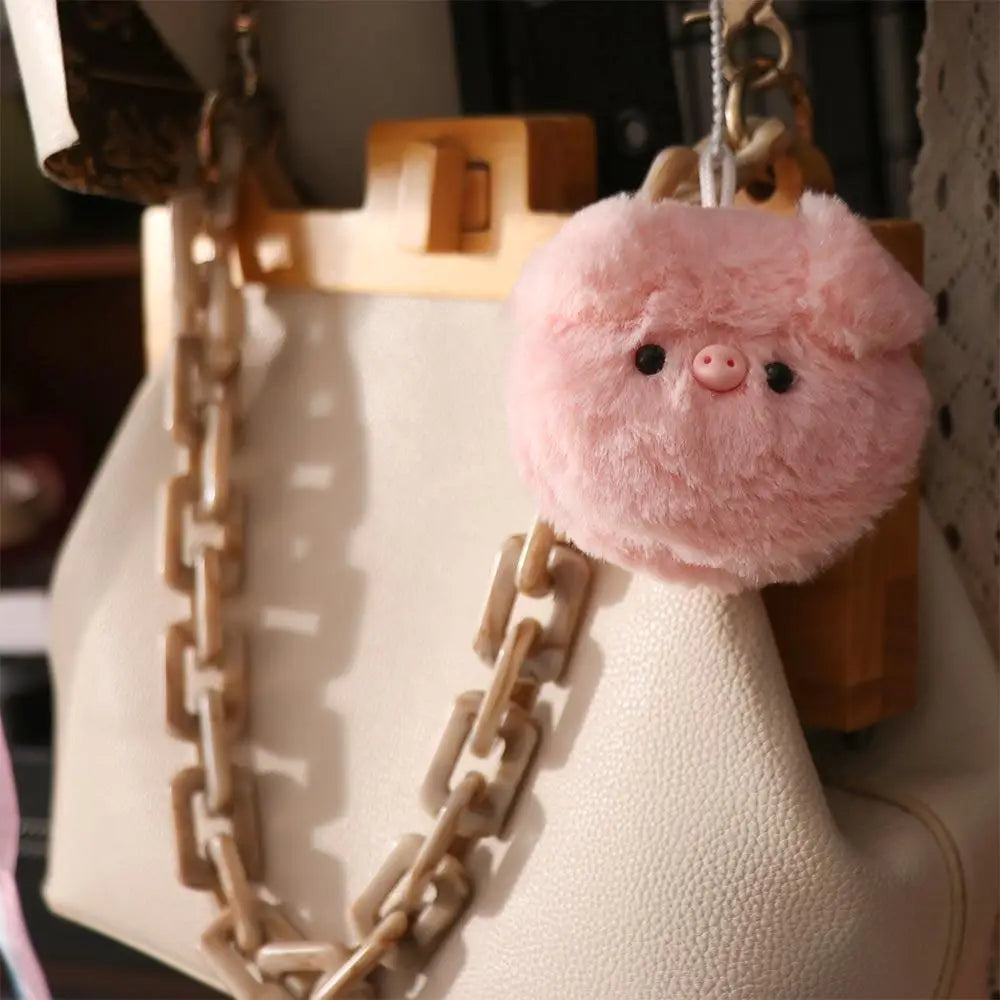 Pink plush kawaii pig keychain hanging on a beige handbag with a chain detail.