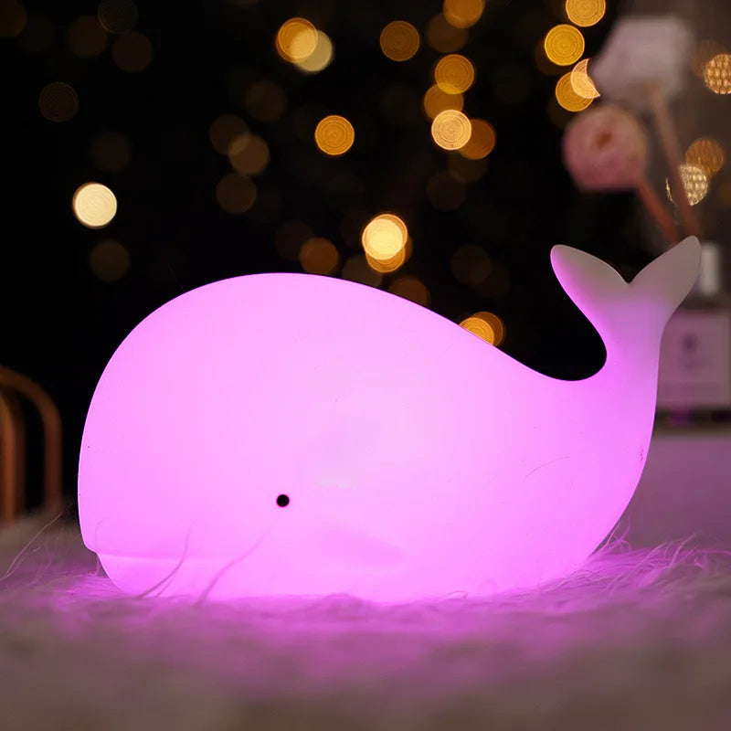 Whale-shaped kawaii night light glowing pink on a soft surface with blurred lights in the background