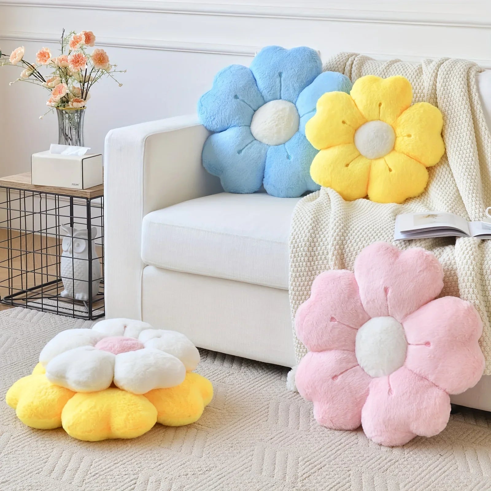 Flower-shaped kawaii cushions in blue, yellow, and pink on a white couch with a side table and flowers in the background.