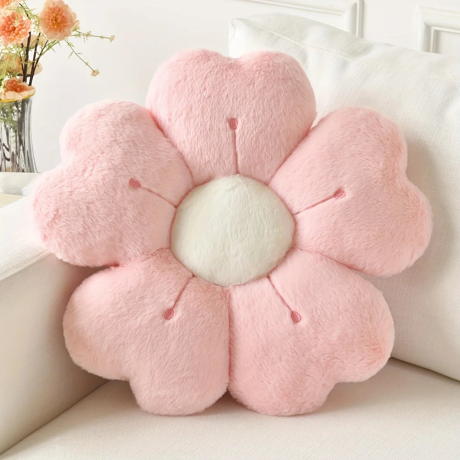 Pink flower-shaped kawaii cushion on a white couch with a blurred floral arrangement in the background.