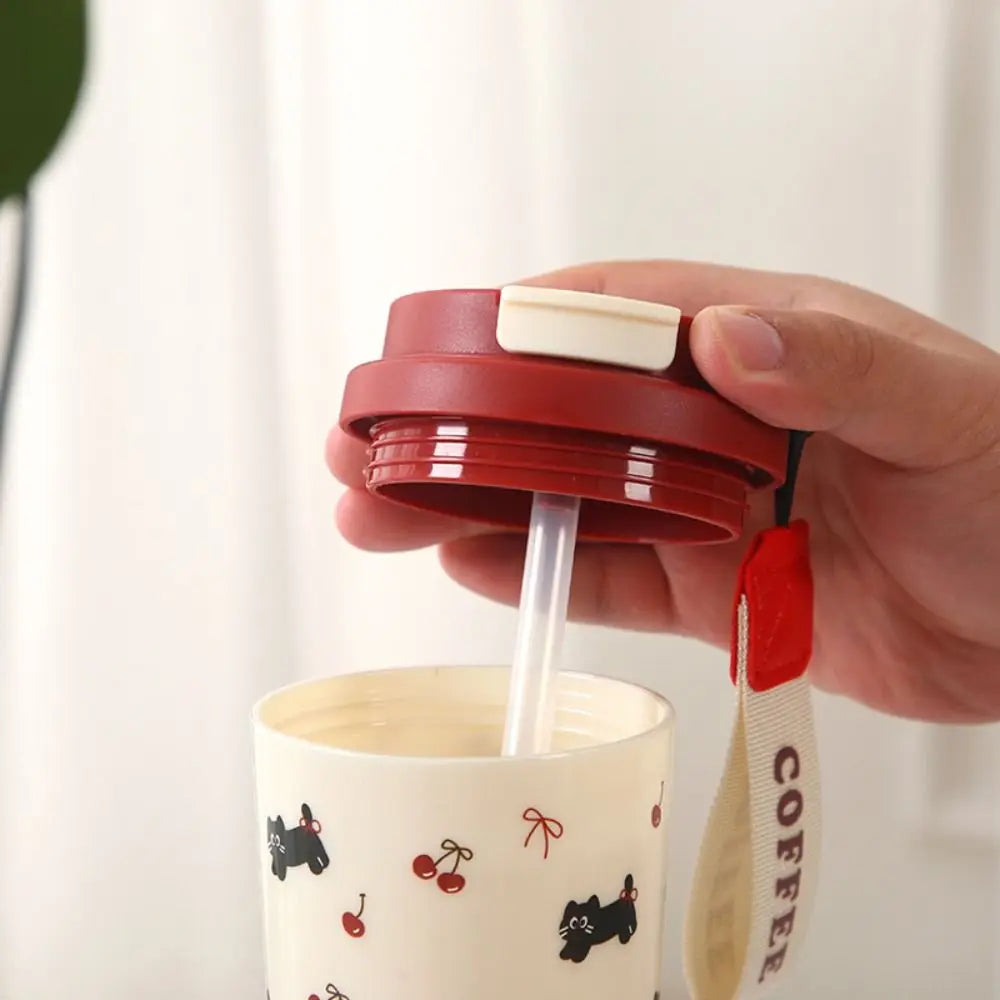 Hand holding a red lid with a straw over a white Kawaii mug with black cat designs on a light background