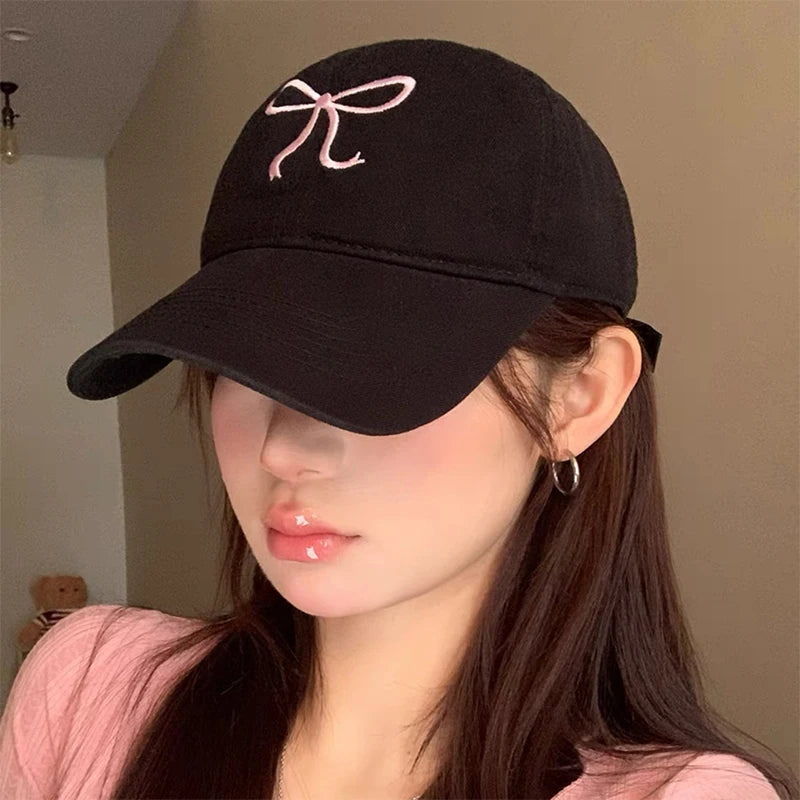 Black Kawaii cap with pink bow design worn by a person indoors.
