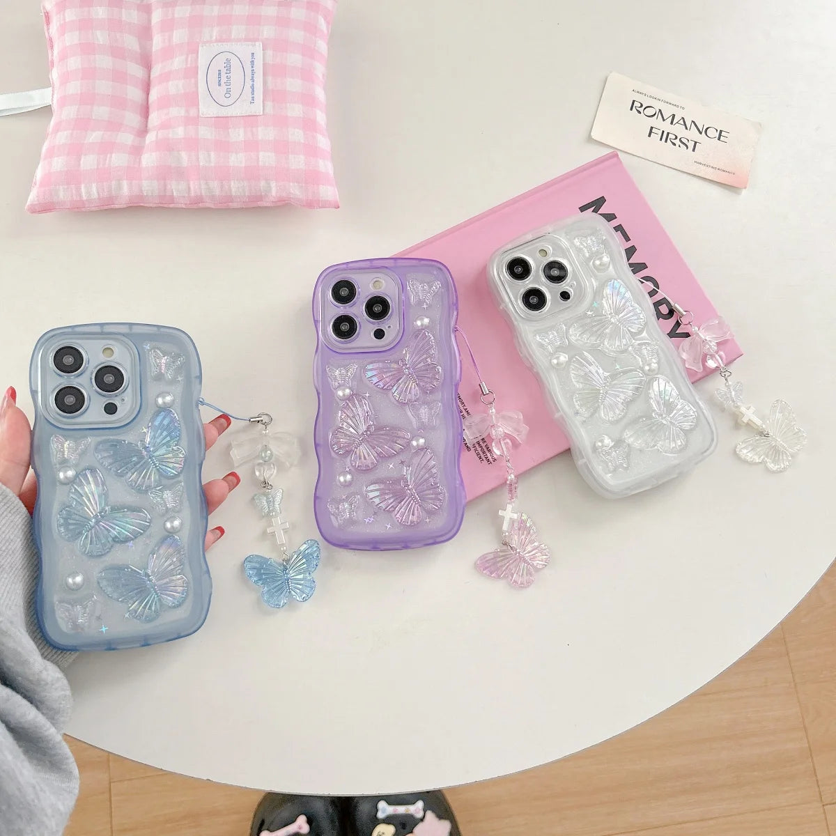 Three kawaii phone cases with butterfly designs on a table with a pink checkered bag and text card in the background.