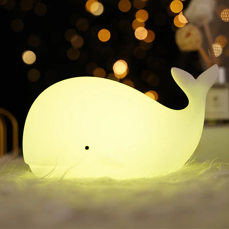 Glowing whale-shaped kawaii night light on a soft surface with blurred lights in the background