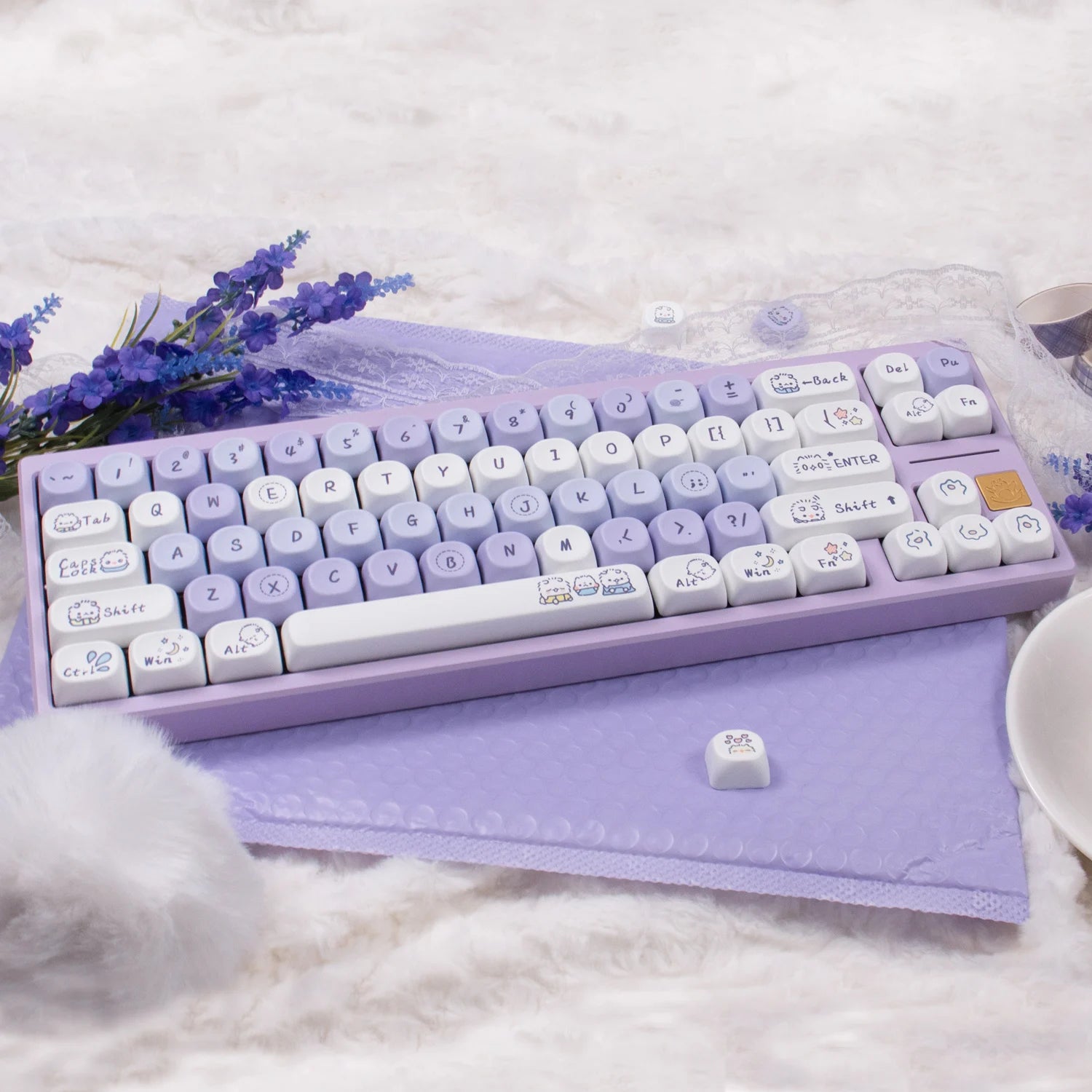 Purple and white Kawaii keyboard with cute keycaps on a soft surface with flowers and a cup.