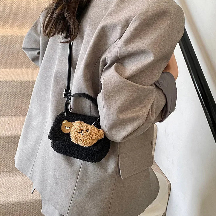 Person holding a small black bag with a Kawaii teddy bear charm, wearing a gray coat.